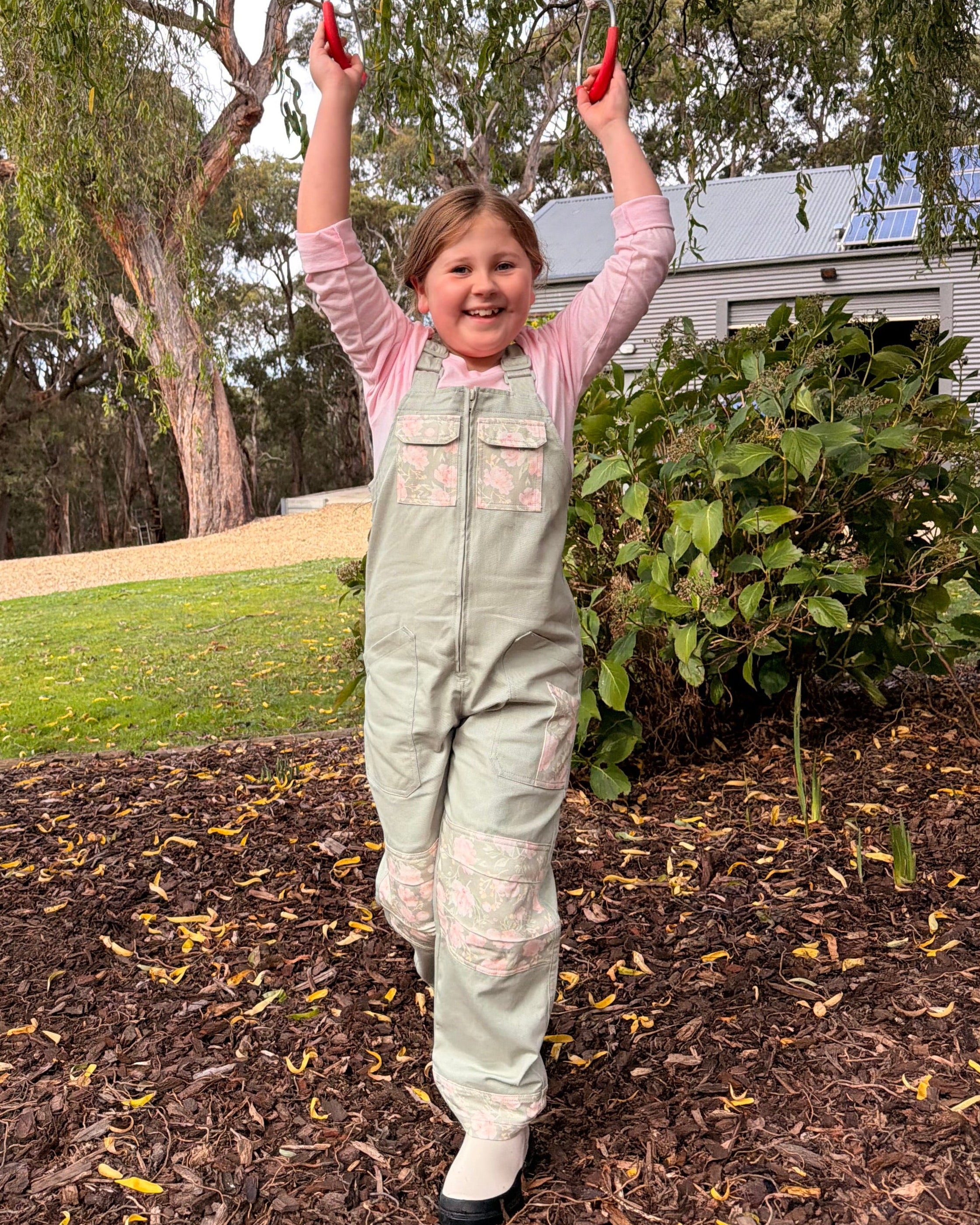 Girl in Lil' Patch Dungarees hanging from a tree swing, arms raised