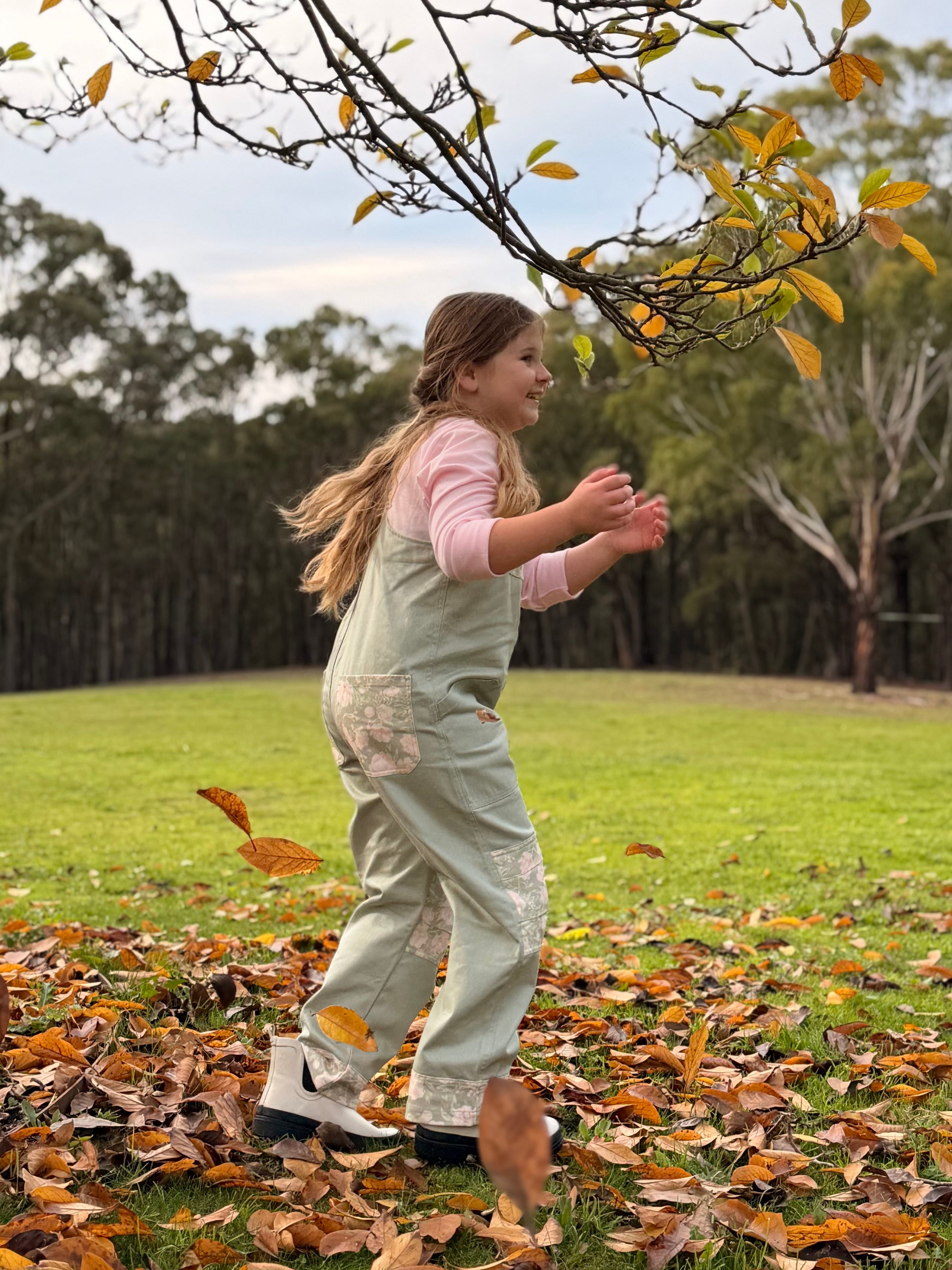 Girl in Lil' Patch Dungarees running through fallen autumn leaves