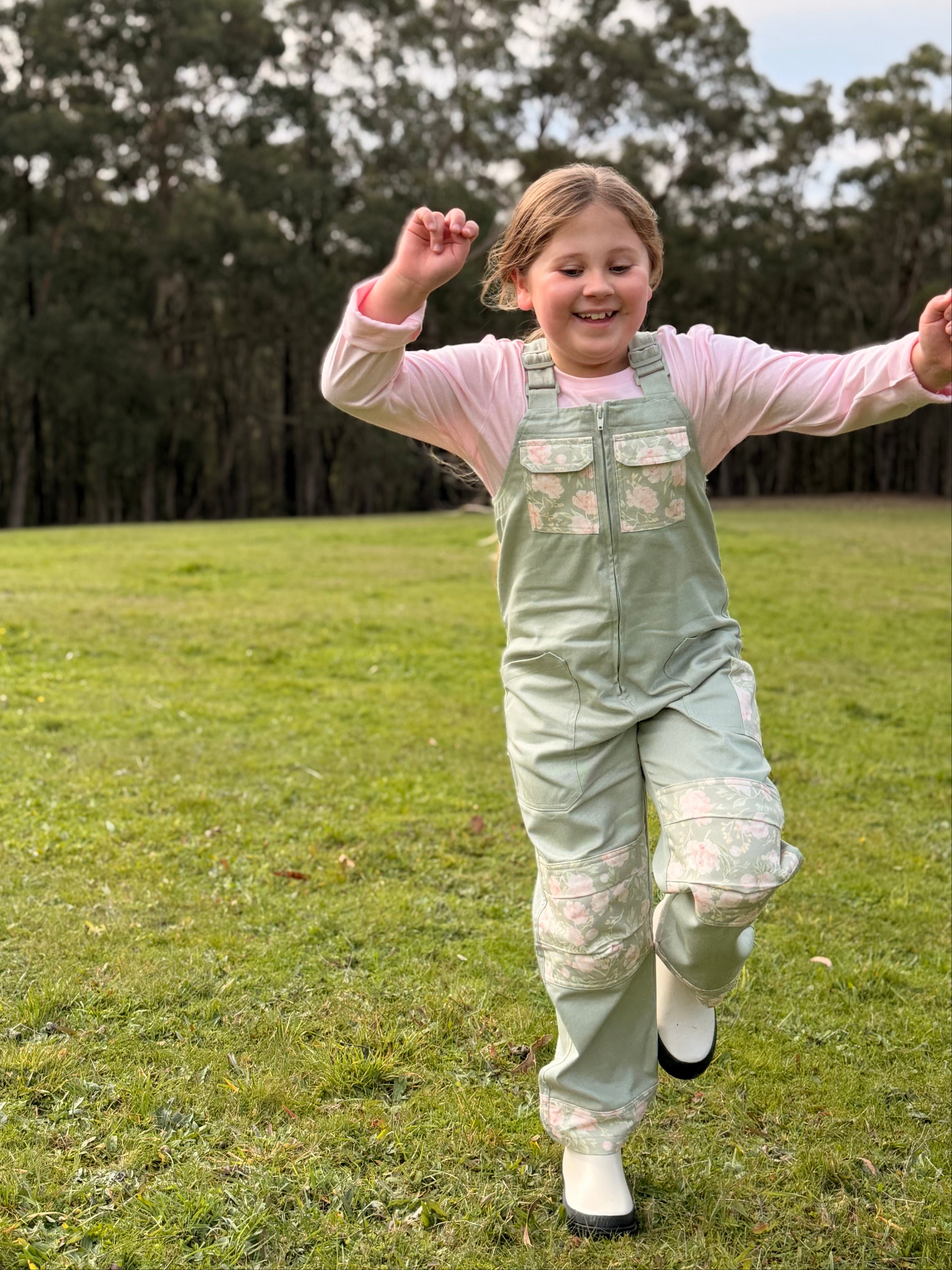 Girl in sage green Lil' Patch Dungarees running on one leg in the grass