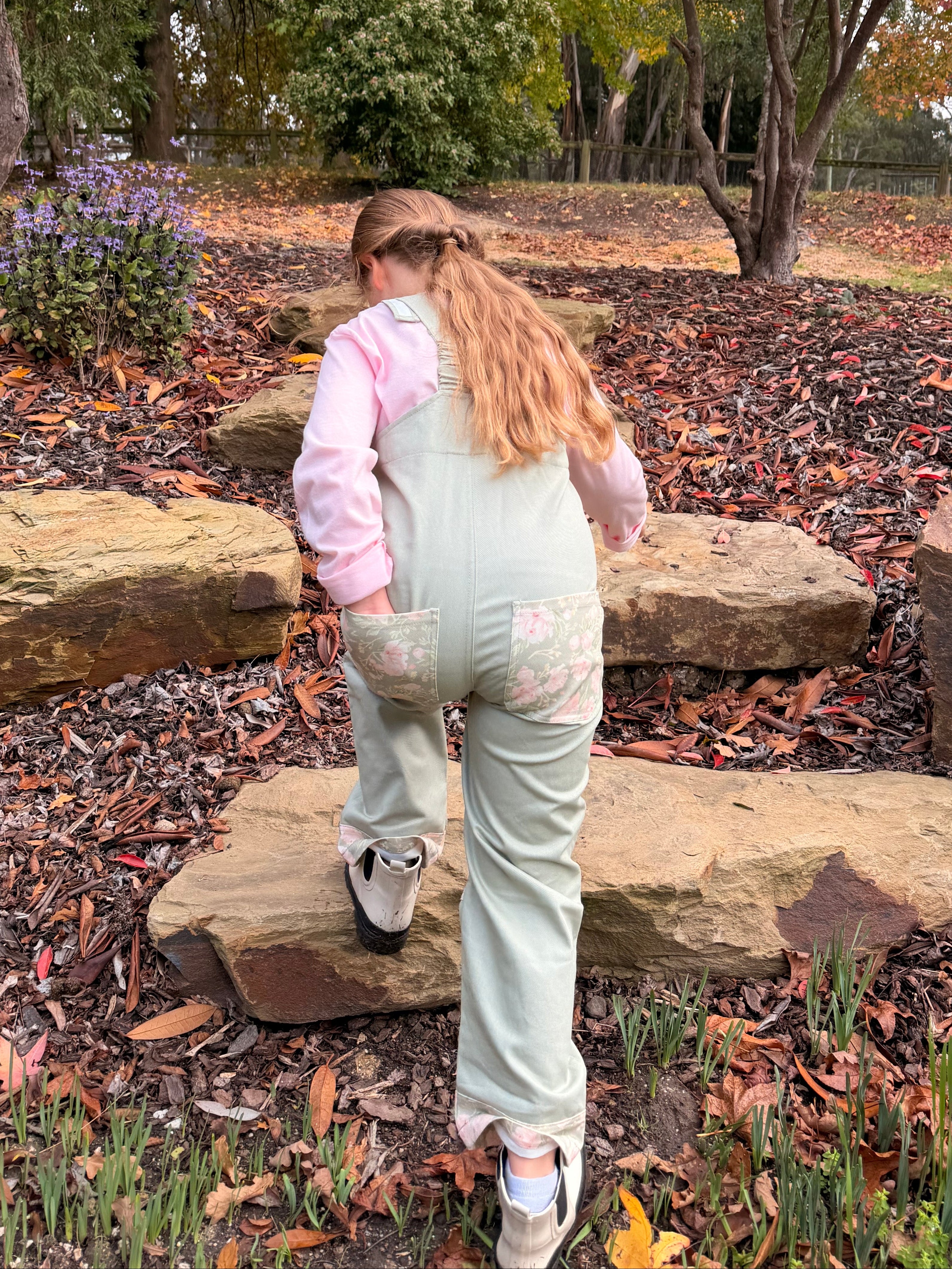 Back view of girl in Lil' Patch Dungarees climbing garden rock steps, showing the floral back pocket