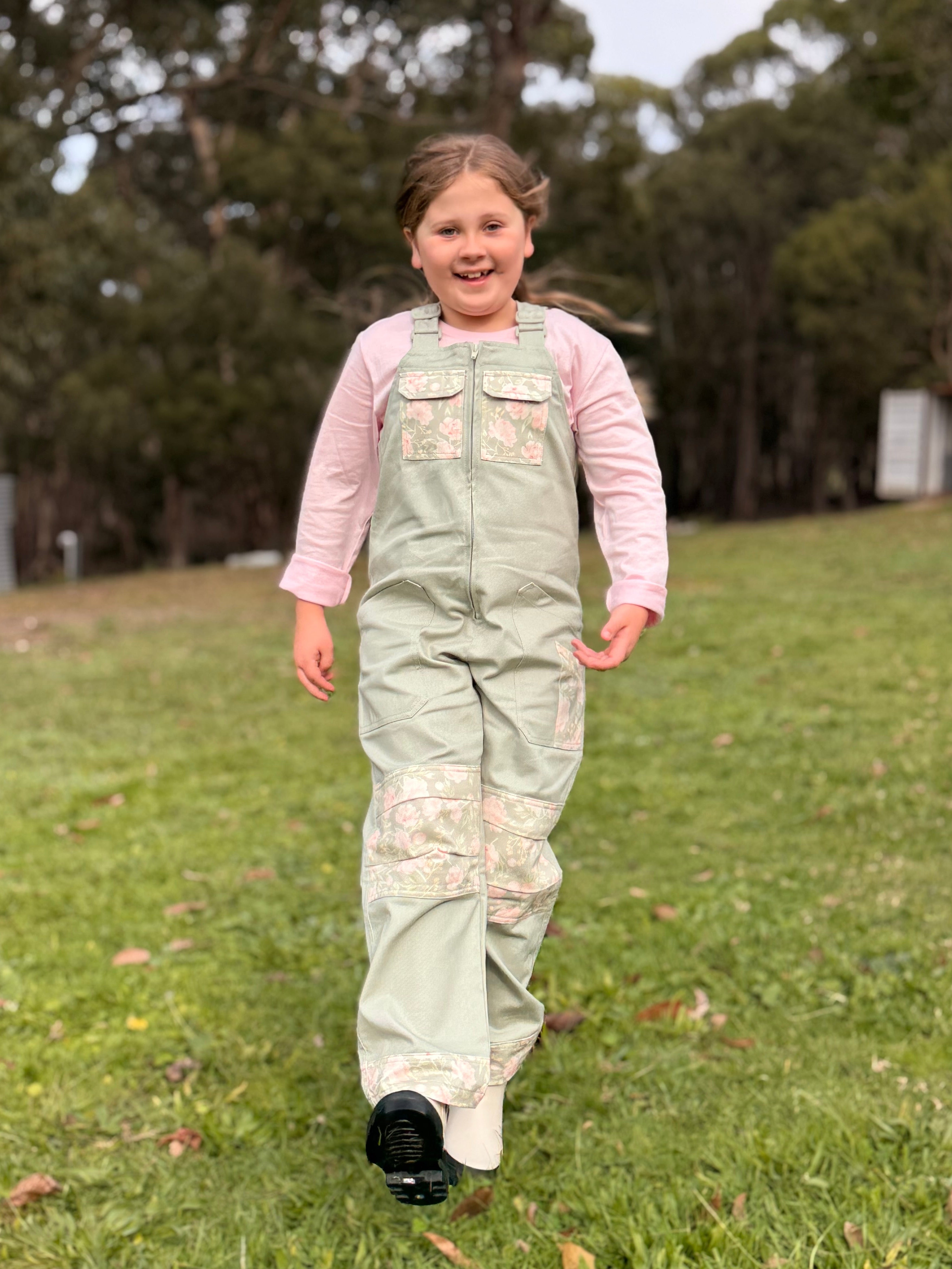 Girl in Lil' Patch Dungarees walking across the grass, smiling
