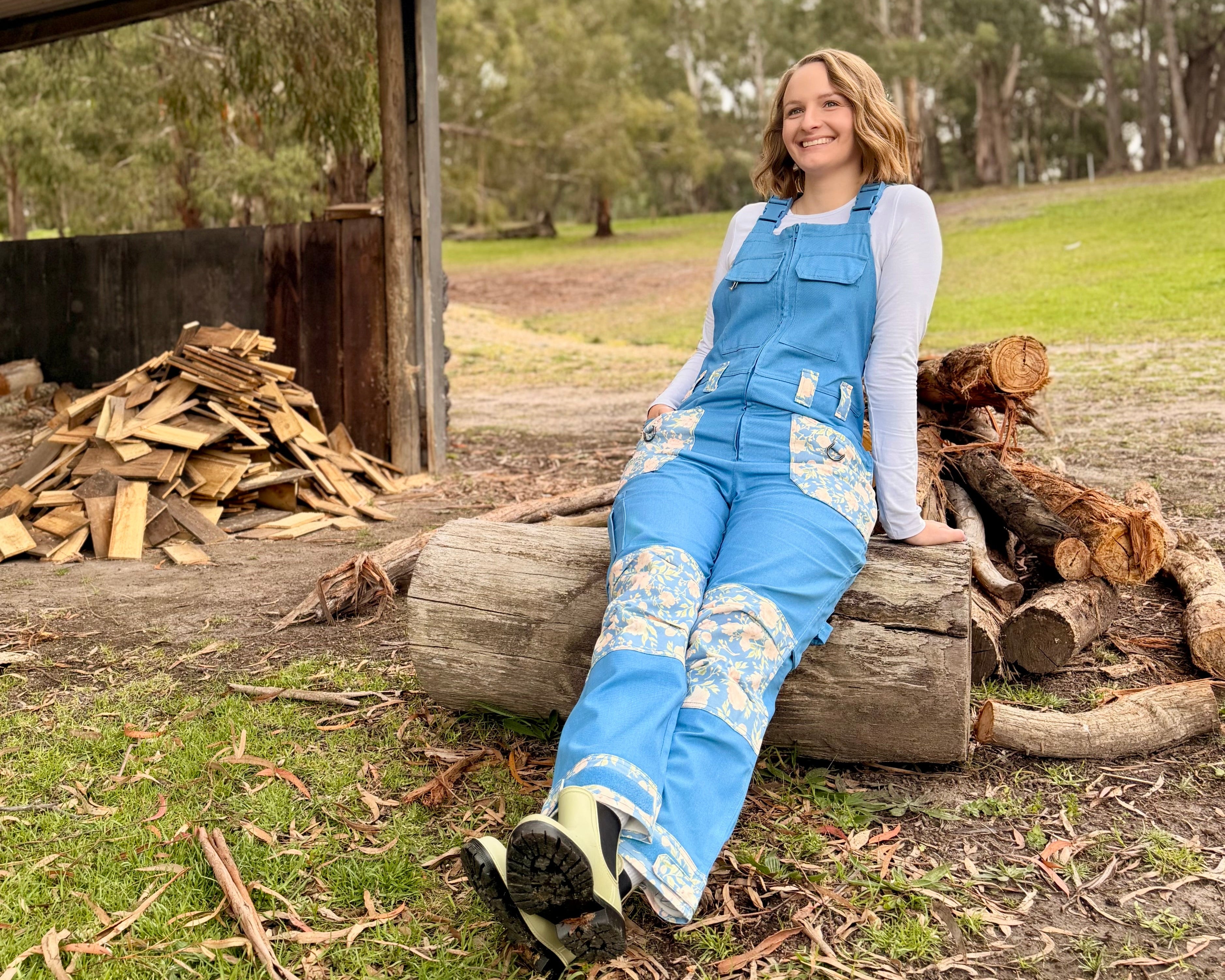 Woman sitting on a log wearing Peony Drop Dungarees, near a woodshed and firewood pile