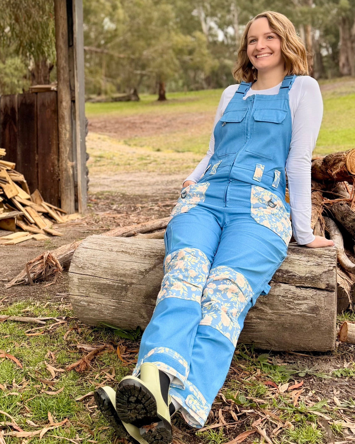 Woman in blue overalls sitting on a log in a rustic outdoor setting with trees and wooden structures.