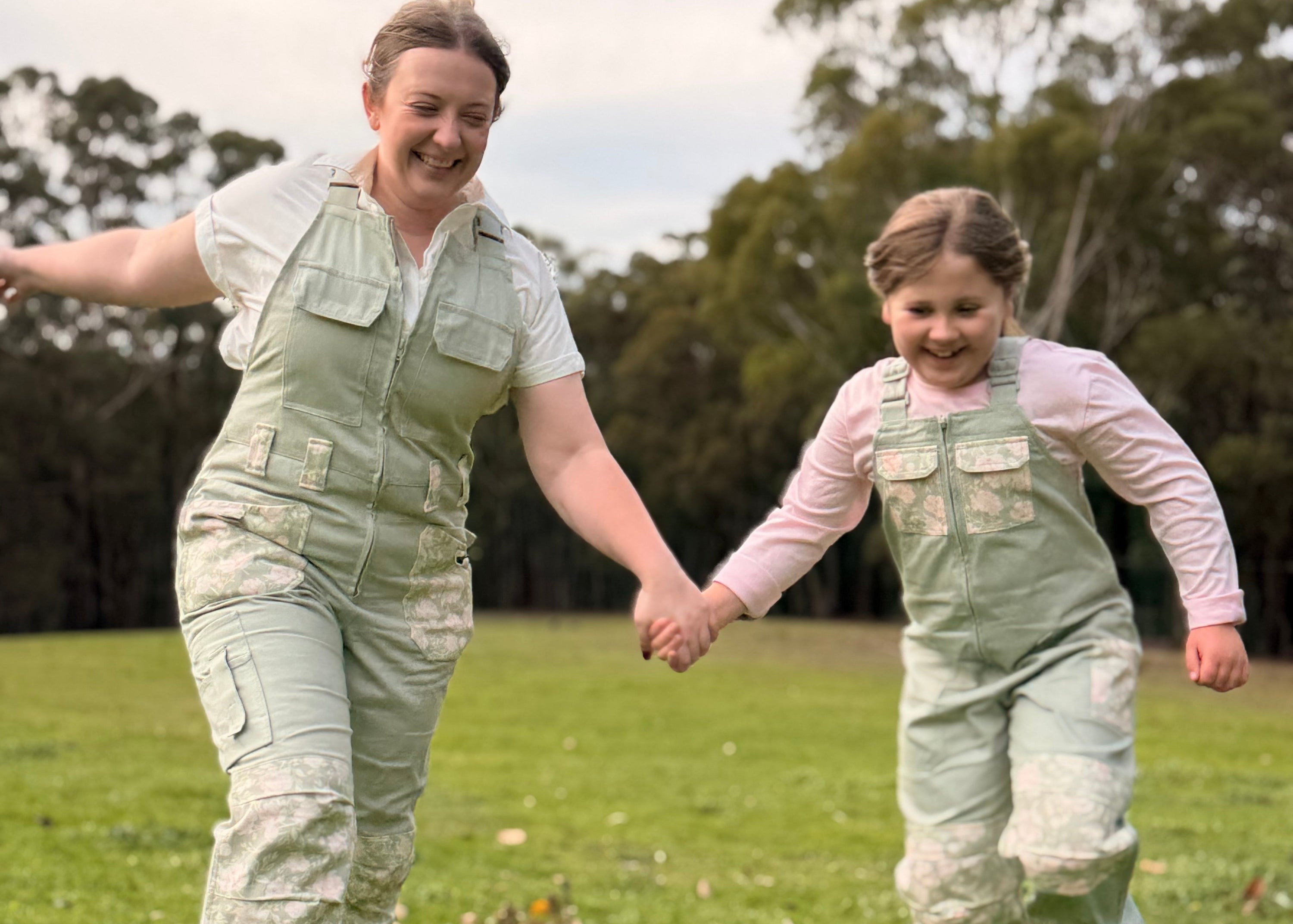 A woman is wearing green overalls while smiling and holding hands with a young girl who is wearing matching green overalls.
