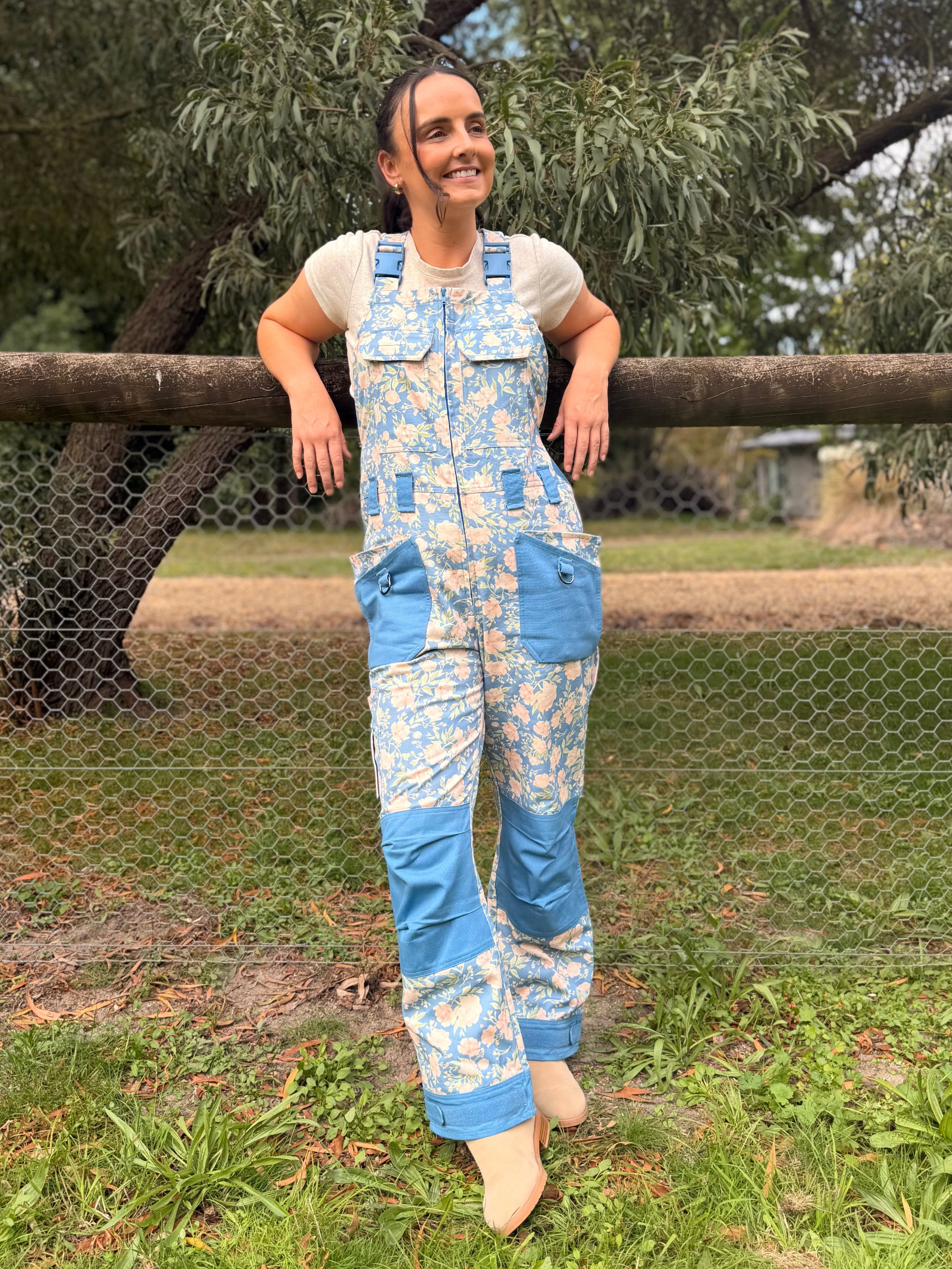 Woman wearing blue floral Peony Mist Dungarees leaning against a wooden fence rail