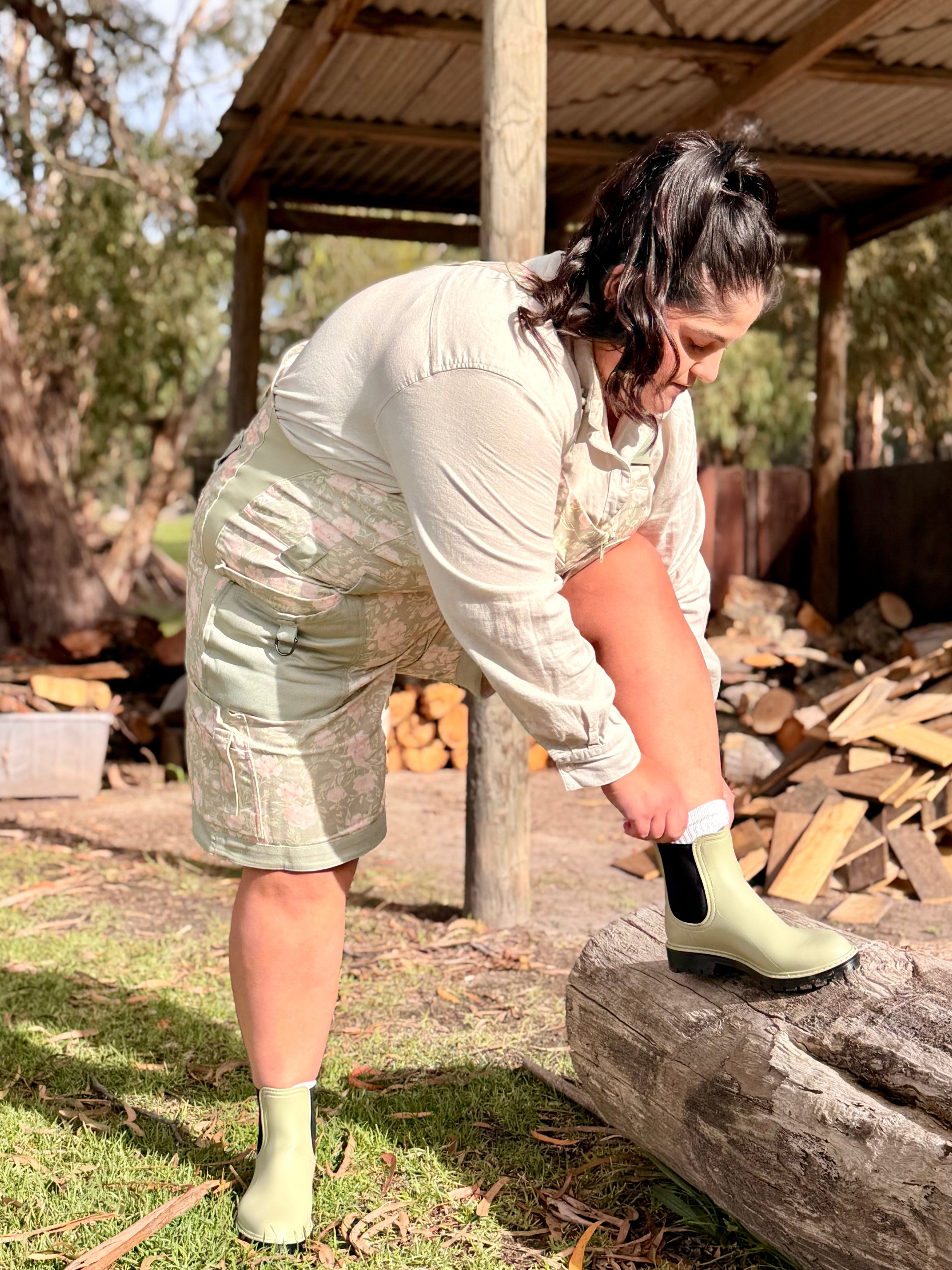 Woman in Peony Meadow Shortalls putting on gumboots near a firewood pile