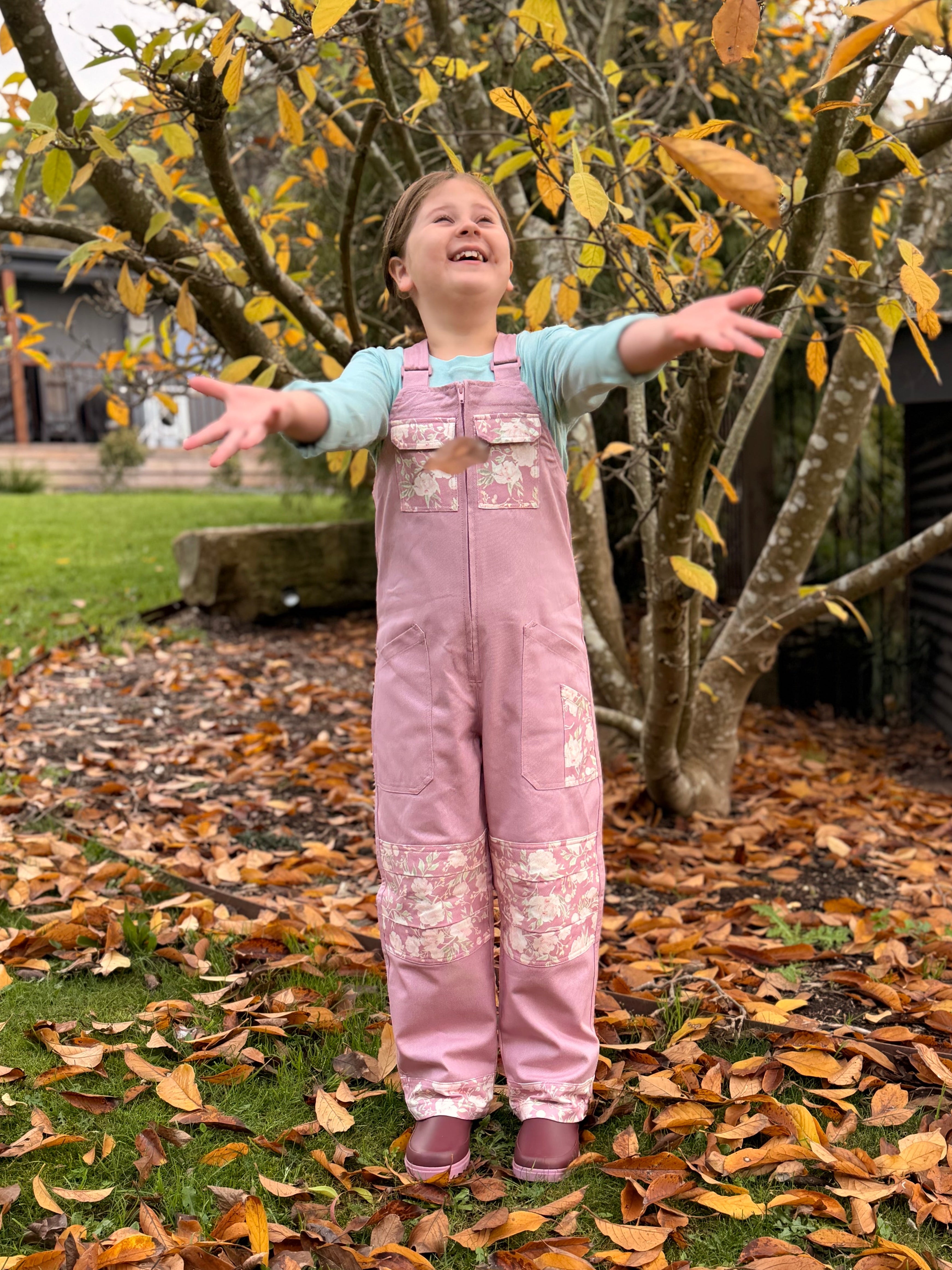 Girl in pink floral Lil' Posy Dungarees standing in autumn leaves with arms outstretched